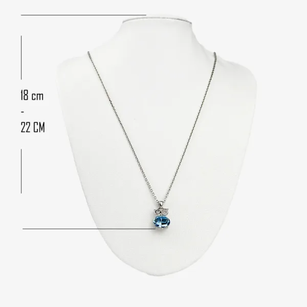 A silver chain necklace with an oval blue crystal pendant and two smaller clear crystal accents, displayed on a white mannequin bust. The necklace is adjustable between 18 cm and 22 cm.