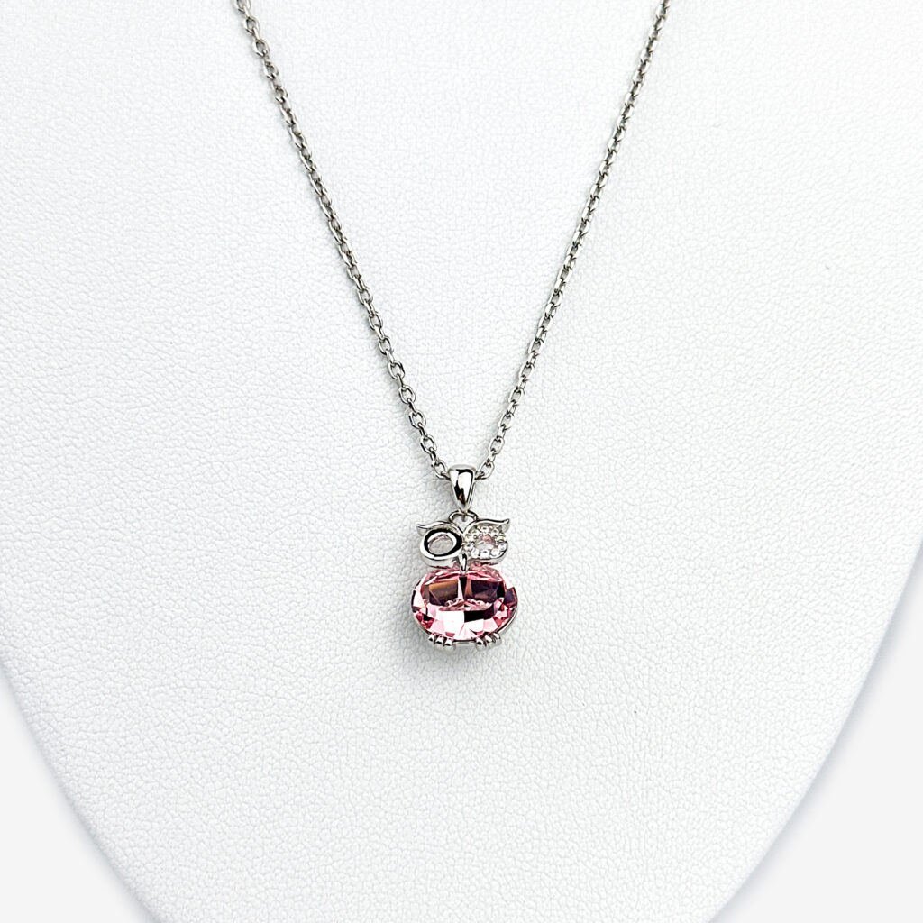 A silver necklace with a pink owl pendant adorned with clear gemstones, displayed on a white mannequin bust.