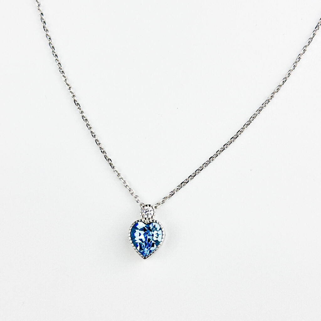 A silver chain necklace with a heart-shaped blue topaz pendant and a small diamond accent at the top, displayed against a white background.