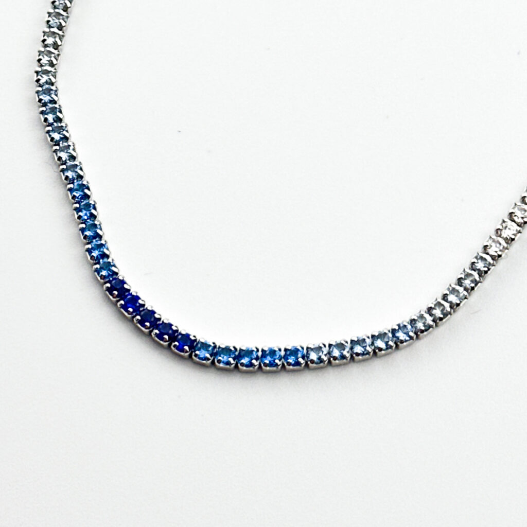 A silver tennis necklace with a gradient of blue sapphire gemstones, transitioning from light to dark blue.