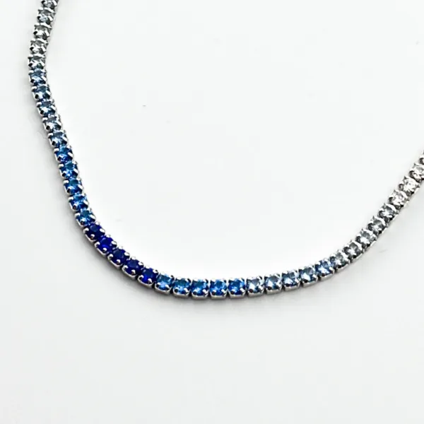 A silver tennis necklace with a gradient of blue sapphire gemstones, transitioning from light to dark blue.