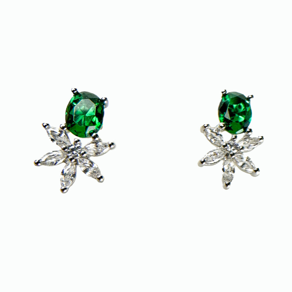 A pair of silver stud earrings with a green oval gemstone set above a cluster of clear gemstones arranged in a floral pattern.